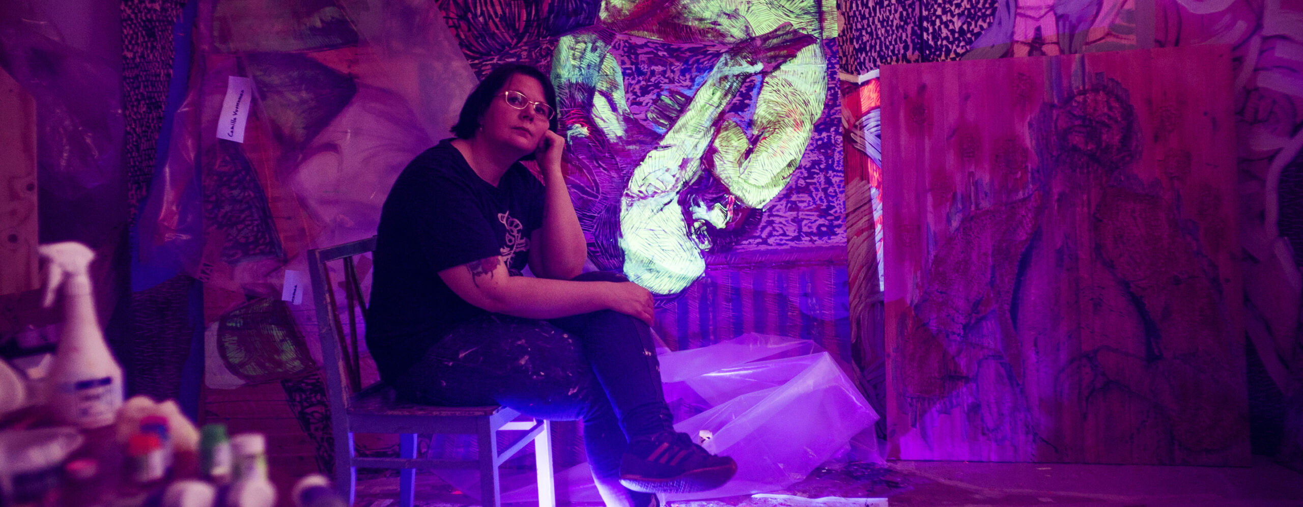 Woman sitting on a chair surrounded by paintings. The lighting is dark fluorescent purple.