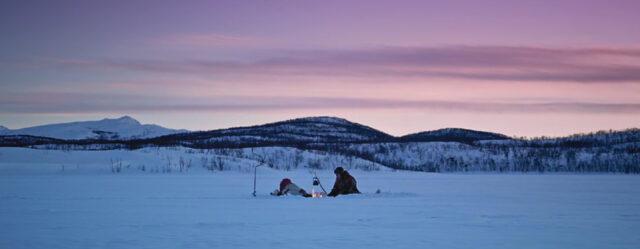Still from the film Giitu showing a man in front of a fire in a snowy landscape with a purple sky in the background